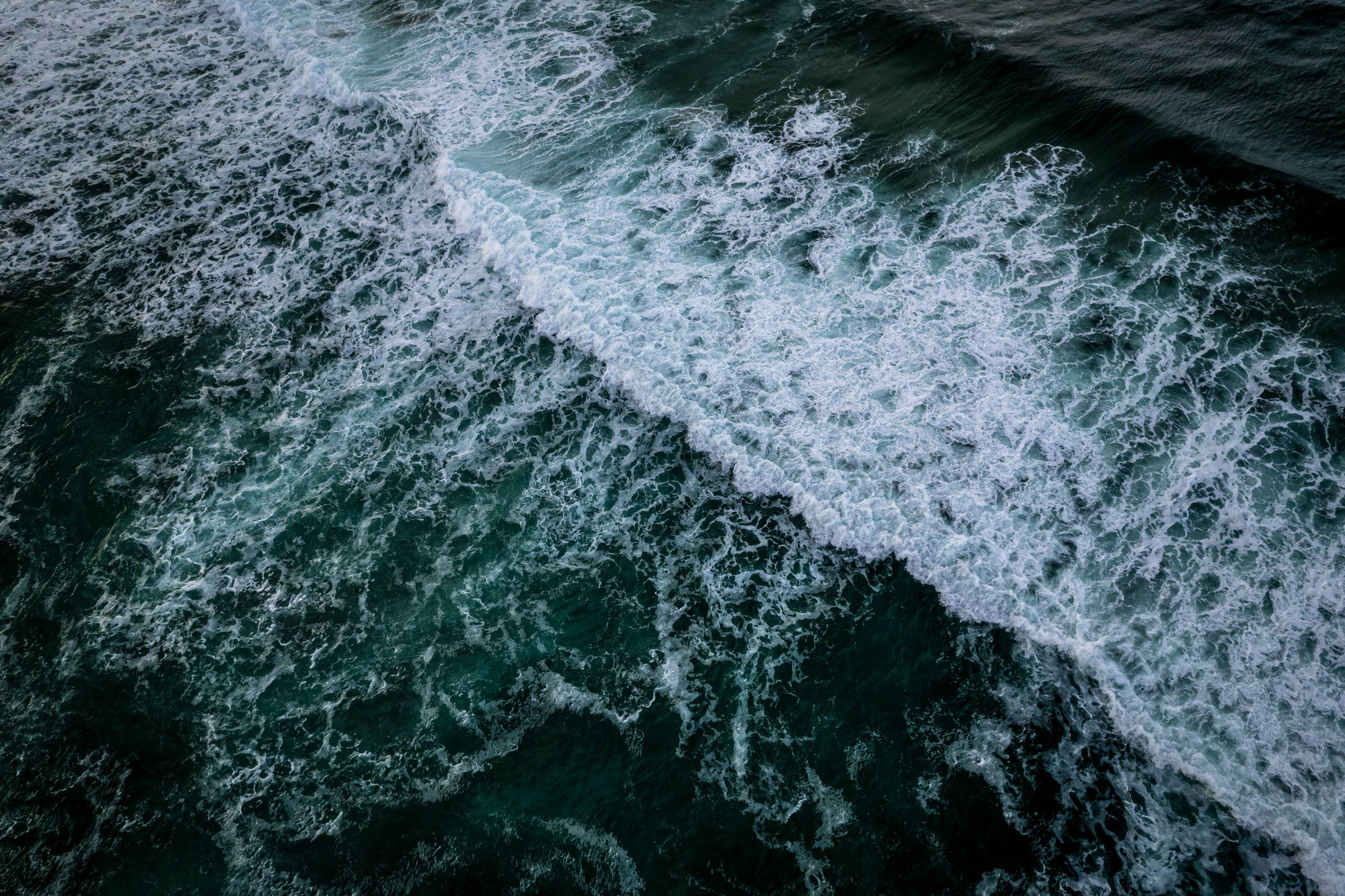 Aerial view of a breaking ocean wave with white foam against dark turquoise-green water.