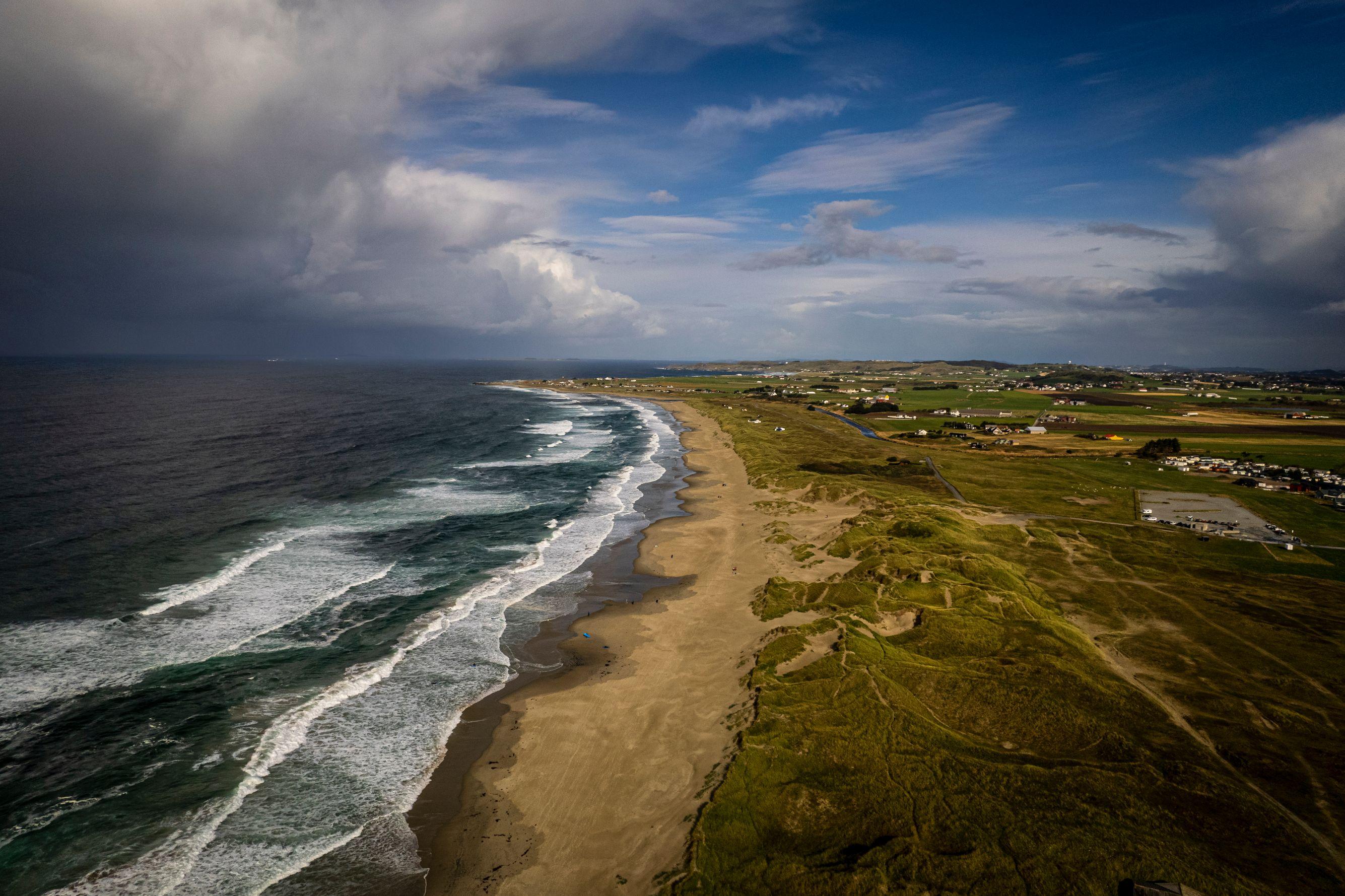Aerial view of a long sandy beach curving along a coastline, with a dramatic stormy sky over the ocean and green farmland inland.
