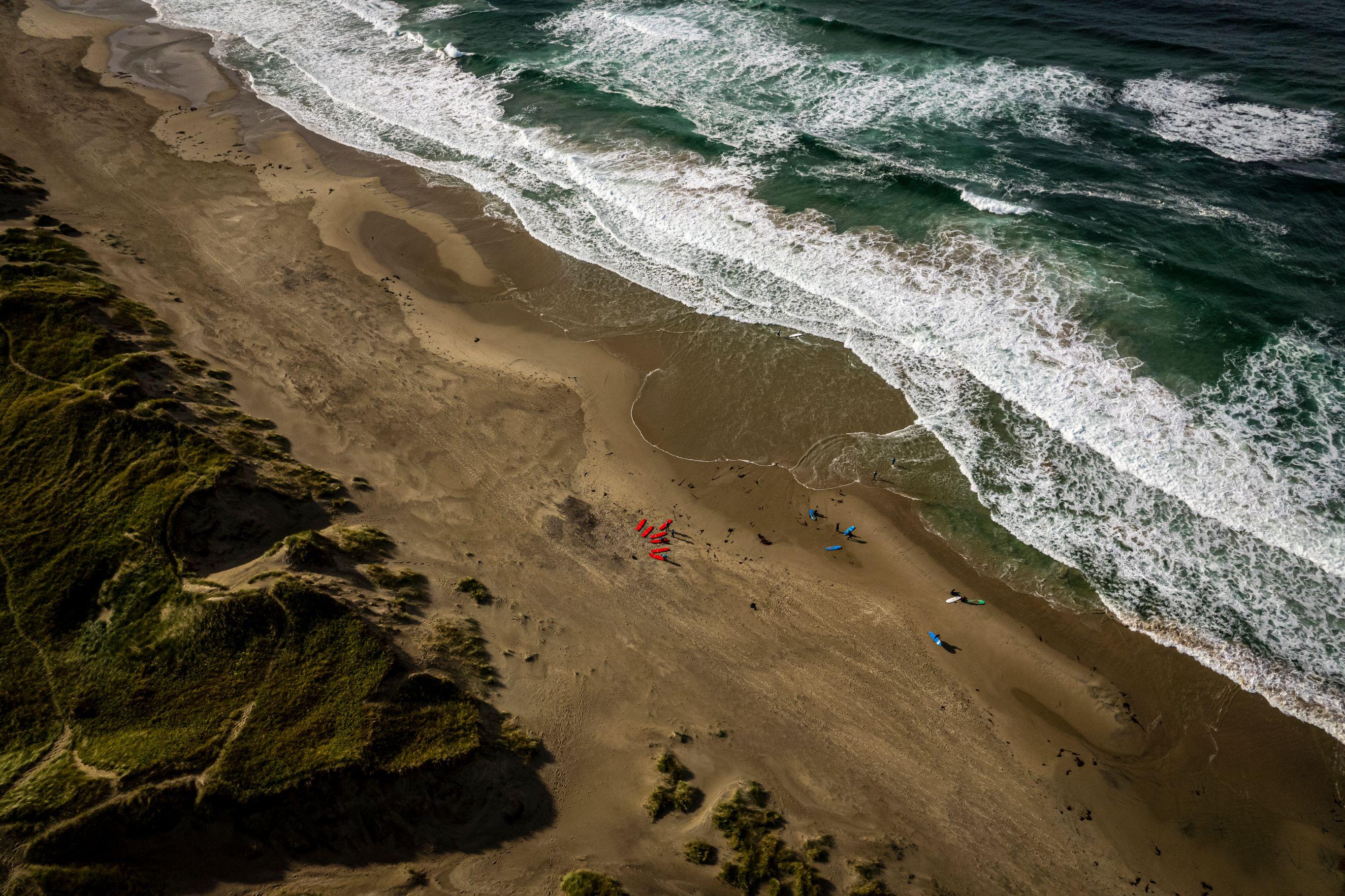 Aerial view of a sandy beach with surfers and colourful surfboards beside breaking ocean waves and a grassy dune.