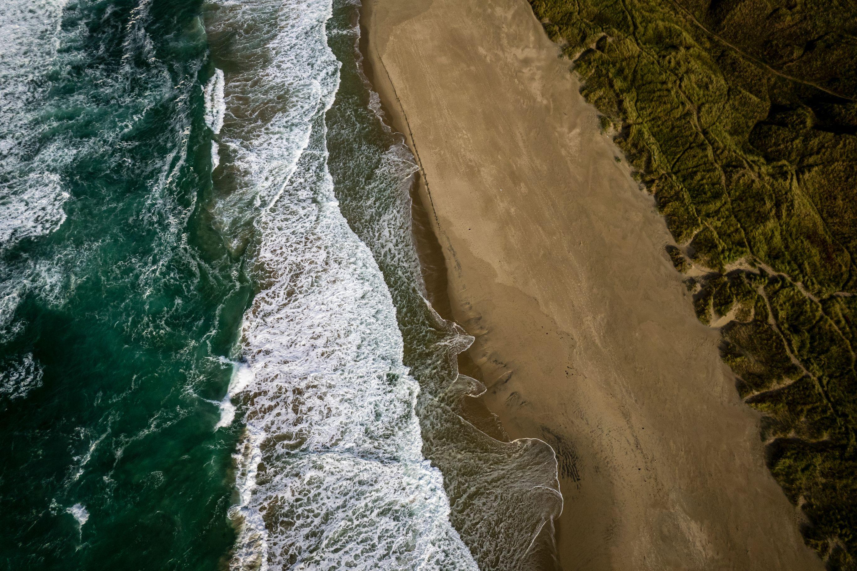 Aerial view of waves breaking onto a sandy beach bordered by green vegetation.